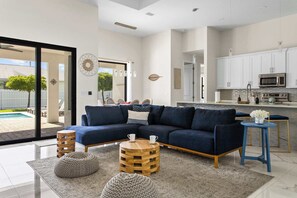 Ein modernes Wohnzimmer mit Eckcouch, rundem Couchtisch und Hocker, eine Küche mit weißen Schränken und Edelstahlgeräten sowie eine Glastür, die zu einem Poolbereich führt.