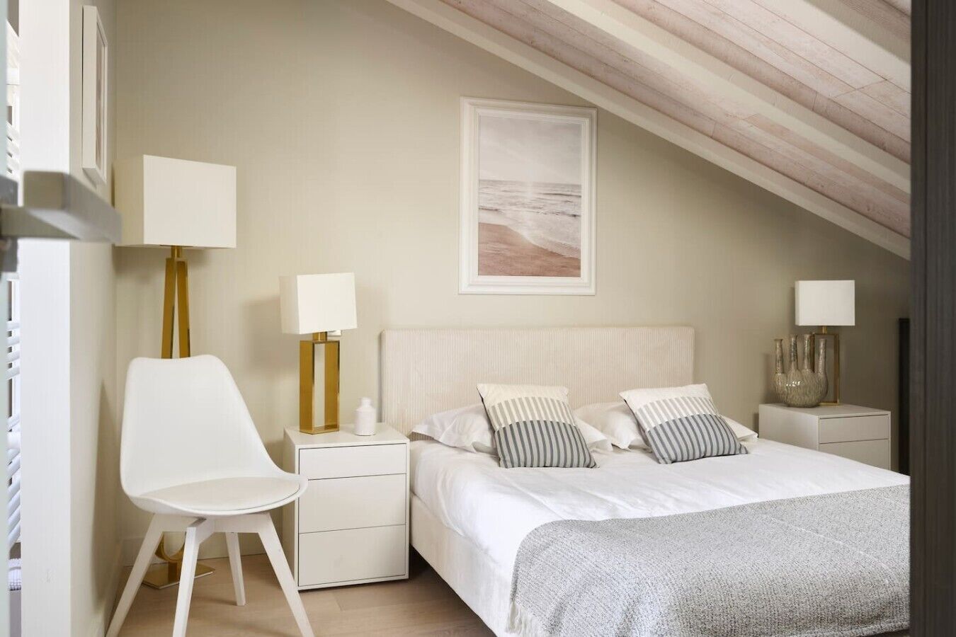 A bedroom with a sloped ceiling, a bed with striped pillows, a white chair, bedside tables with lamps, and a framed picture on the wall.