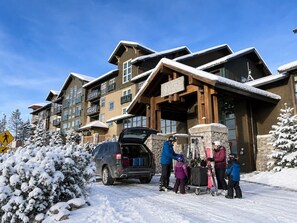Ein mehrstöckiges Gebäude mit Skiurlaubseingang, ein Auto mit Gepäck und schneebedeckte Bäume.