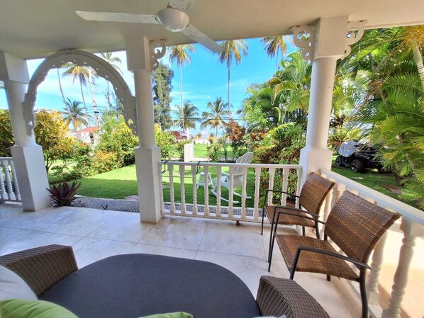 A covered outdoor seating area with a table and chairs, a white railing, and a view of a garden with palm trees. - Rare Sea View Gem Steps from the Village for 4per (Las Terrenas)
