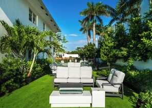 A modern outdoor seating area with a sofa and chairs, surrounded by lush greenery and palm trees.
