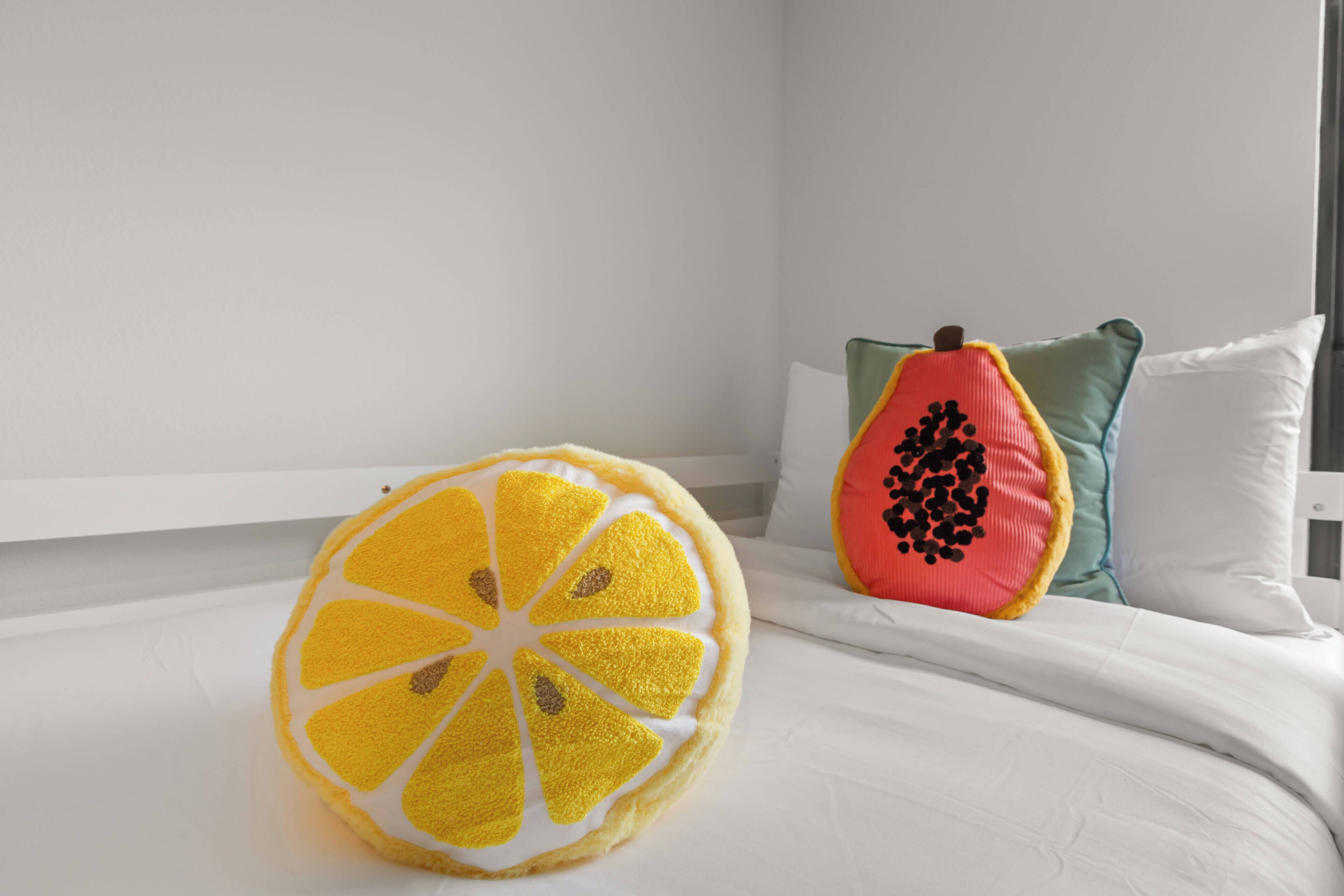 A bed with white bedding, a pillow with a fruit design, and a fruit-shaped pillow.