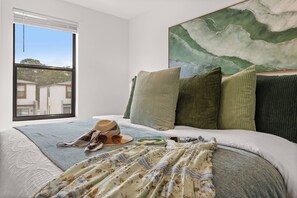 Un dormitorio con un ventanal grande, una cama con muchas almohadas, una colcha y un sombrero sobre la cama.
