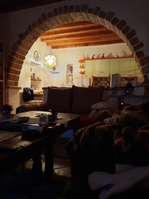 Een gezellige woonkamer met een bakstenen boog die leidt naar een keuken, met een houten tafel, een bank en diverse keukenapparaten.