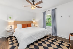 Ein Schlafzimmer mit einem Bett, Nachttischen, einem Deckenventilator, einem Fenster mit Vorhängen und Blick ins Grüne.