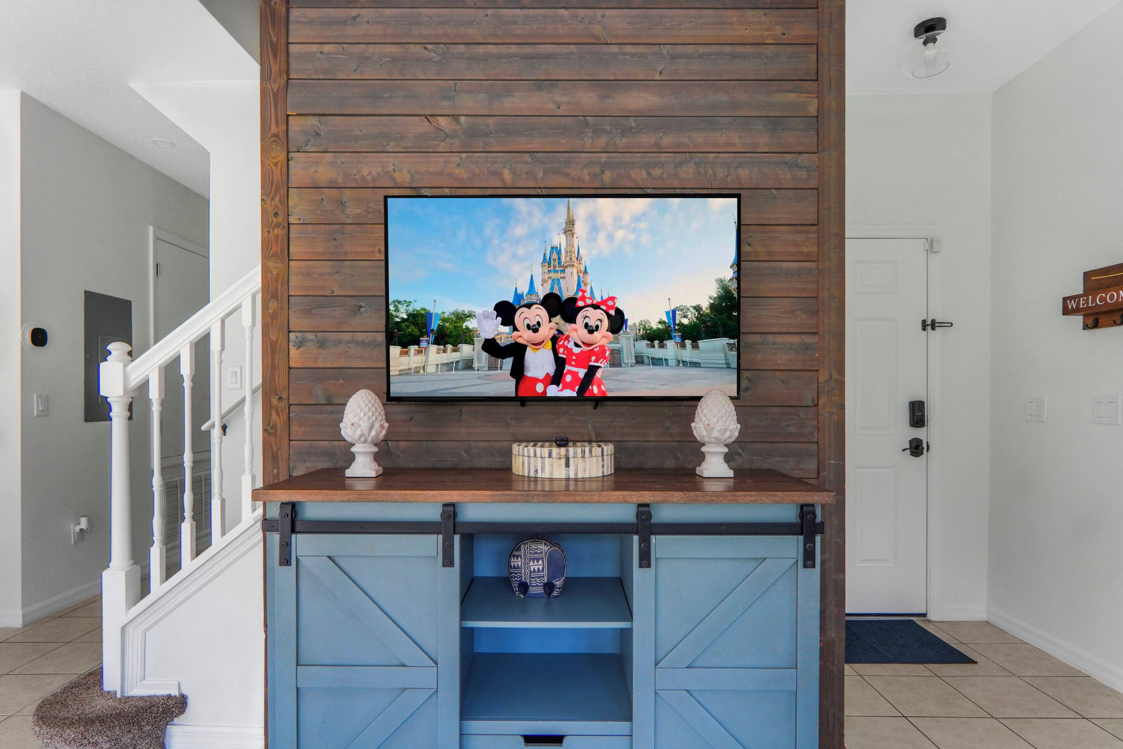 Ein Fernseher an einer Holzwand, der ein Bild mit Disney-Motiv zeigt, ein blauer Unterhaltungsschrank darunter und eine weiße Tür auf der rechten Seite.