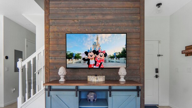 Television on asennettu puiseen seinään, ja sen yläpuolella on Disneyn teemalla koristeltu kuva. Sen alla on sininen viihdekeskus ja oikealla puolella valkoinen ovi.