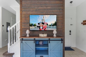 A television mounted on a wooden wall with a Disney-themed picture, a blue entertainment center below, and a white door to the right. - Encantada 4BR Private Pool Near Disney (Kissimmee)