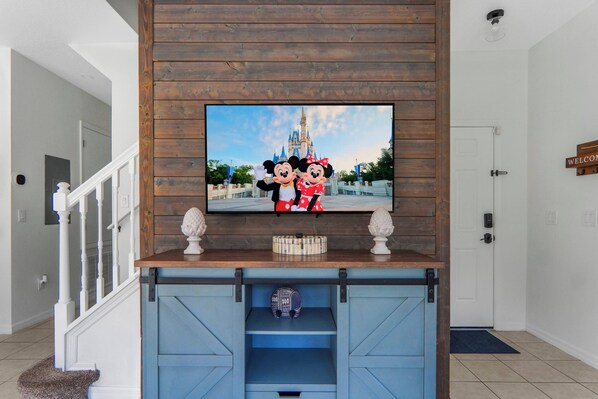 A television mounted on a wooden wall with a Disney-themed picture, a blue entertainment center below, and a white door to the right. - Encantada 4BR Private Pool Near Disney (Kissimmee)