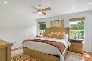 A bedroom with a bed, a ceiling fan, a nightstand with a lamp, and a window with a view of greenery.
