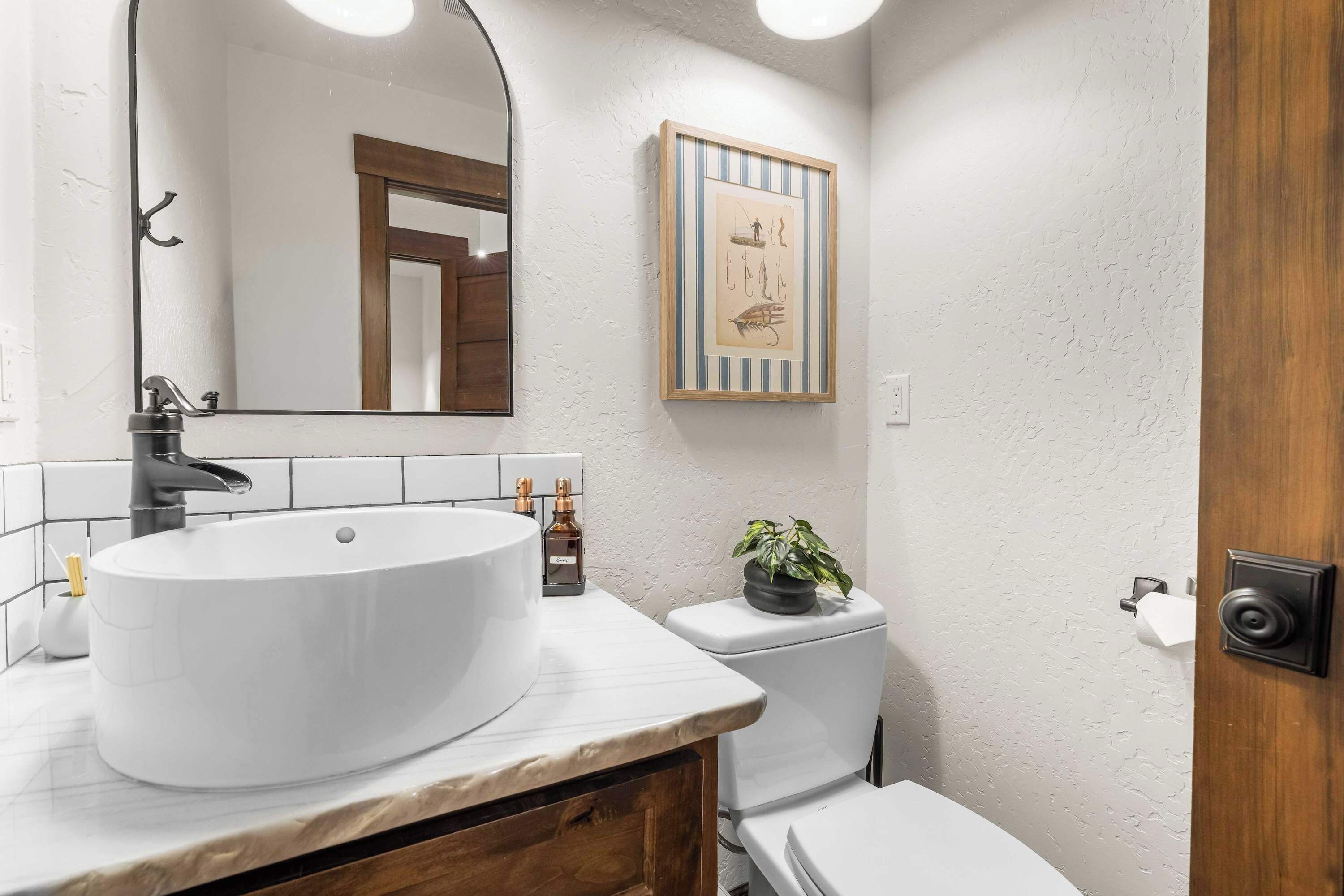 Een badkamer met een grote witte wastafel, een spiegel, een kunstwerk in een lijst, een toilet en een plant in een pot.