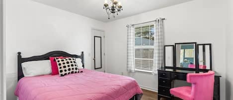 Une chambre à coucher avec un lit, un bureau, une chaise, un miroir et une fenêtre avec des rideaux.
