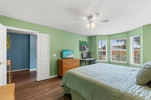 Ein Schlafzimmer mit einem Bett, einem Schreibtisch mit Computer, einem Fernseher und einem Deckenventilator.