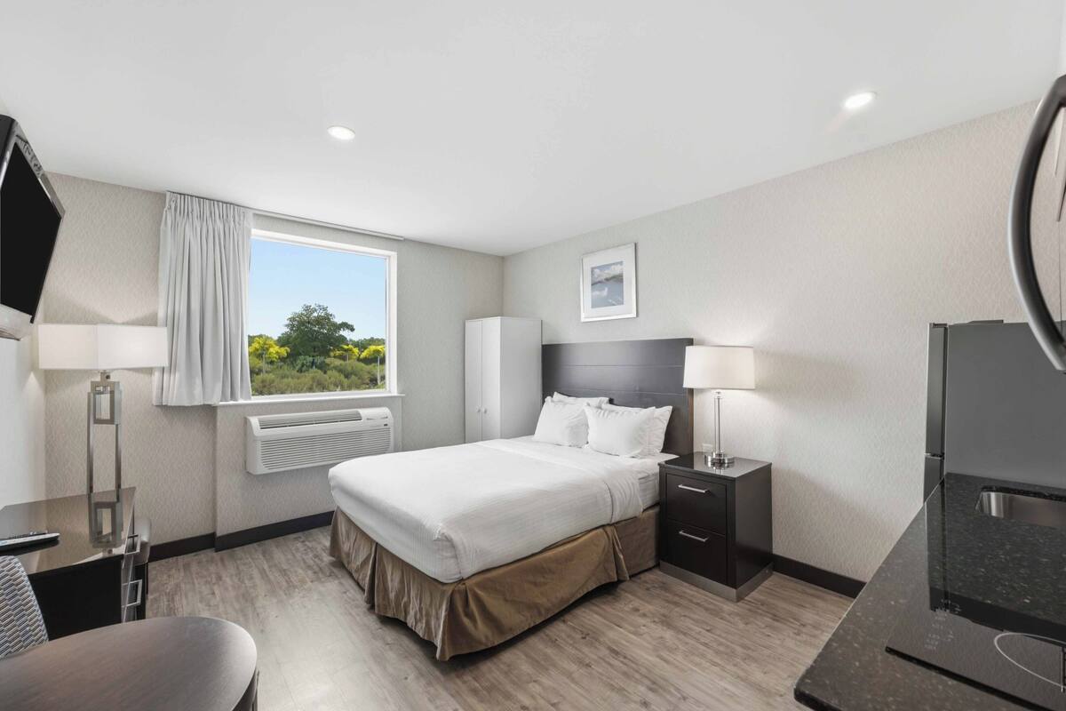 A hotel room with a bed, bedside tables, a desk, a chair, a window with curtains, a picture on the wall, and a refrigerator.
