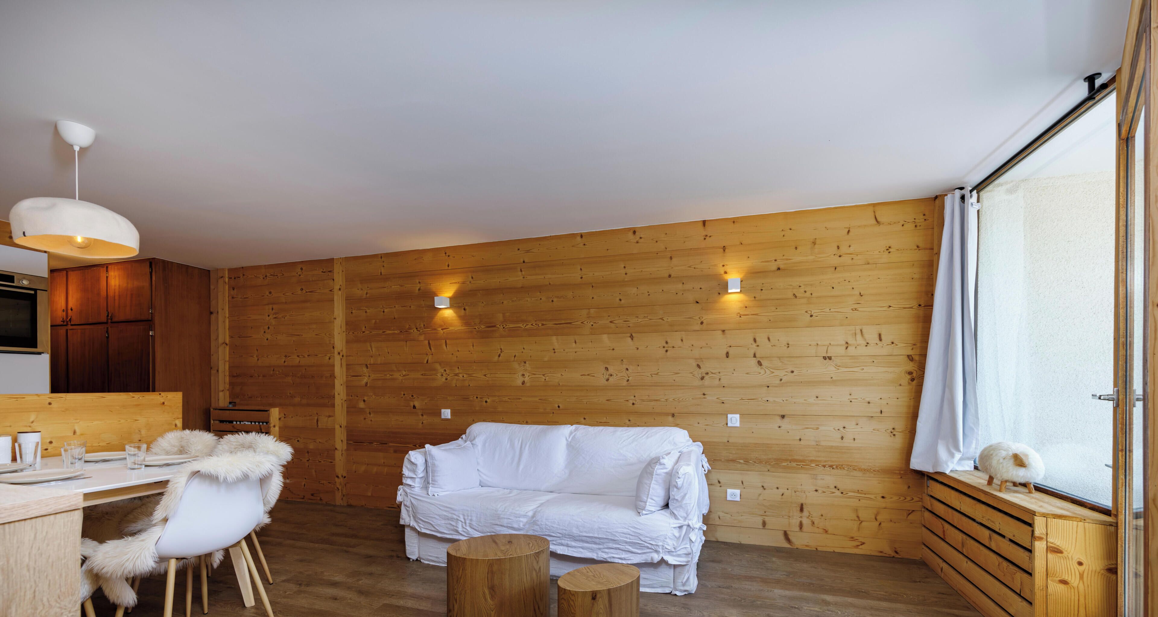 Een gezellige kamer met een houten wand, een witte bank, een eettafel met stoelen en een raam met gordijnen.