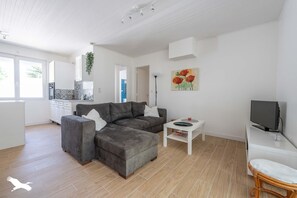 Un salon moderne avec un canapé d’angle gris, une table basse blanche, un téléviseur à écran plat et un fauteuil en osier.