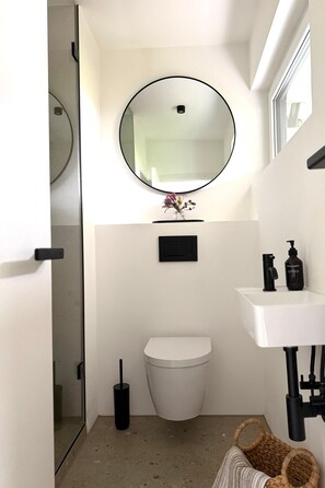 A compact bathroom with a toilet, a round mirror, a small shelf with a vase, and a white sink with a black faucet.
