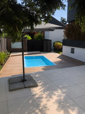 A modern outdoor swimming pool with a wooden deck, surrounded by a white wall and a metal gate.