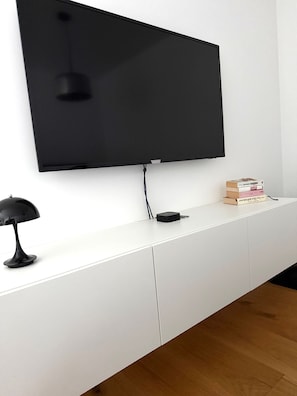 A flat-screen TV mounted on a white wall, a black desk lamp, and a stack of books on a white console table. - Modern House with Private Pool, Garden, Terrace & Wi-Fi in Burgdorf (Burgdorf)