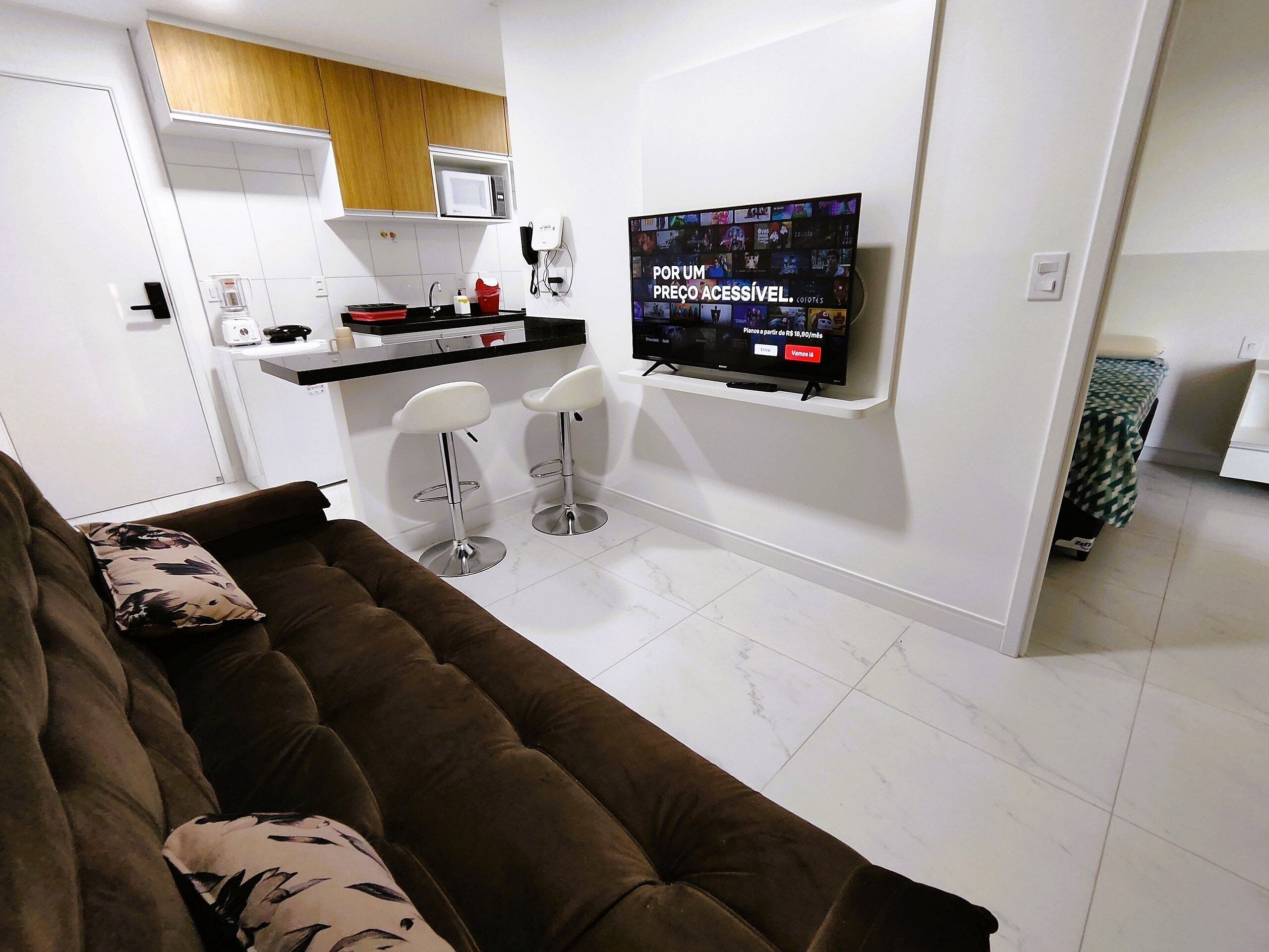 Un salon moderne avec un canapé marron, deux tabourets de bar blancs, un téléviseur à écran plat fixé au mur, un coin cuisine équipé d’un four à micro-ondes et un meuble en bois.