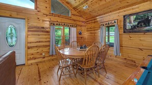 Ein Holzblockhaus-Interieur mit Esstisch, Stühlen, einer Bank, einem Fenster mit Vorhängen und einer Bär-Dekoration.