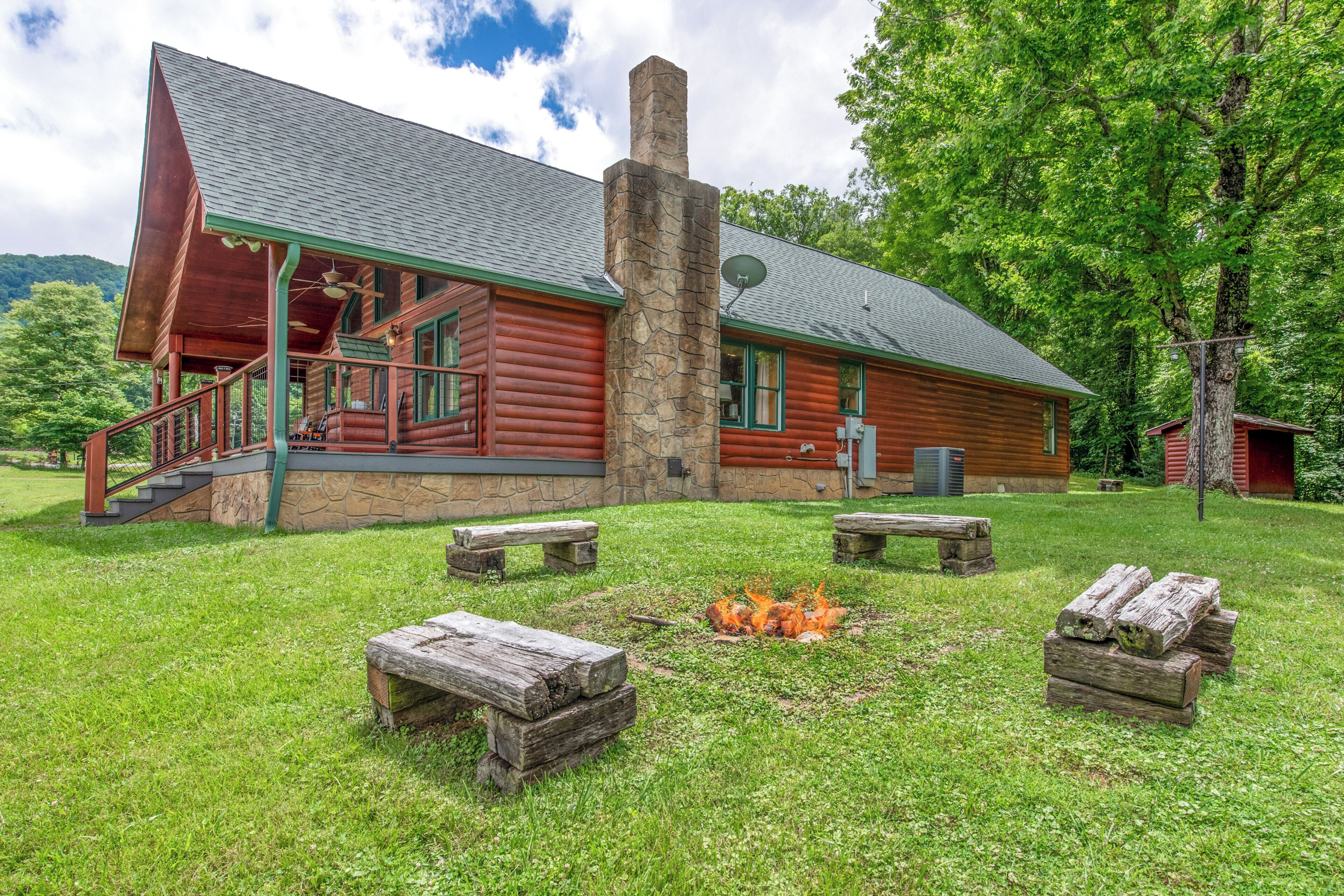 A log cabin with a stone chimney, a porch, and a fire pit surrounded by benches.