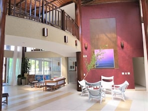 A spacious lobby with a high ceiling, a balcony, a large window, and a seating area with white chairs. - Bech Living (Aquiraz)