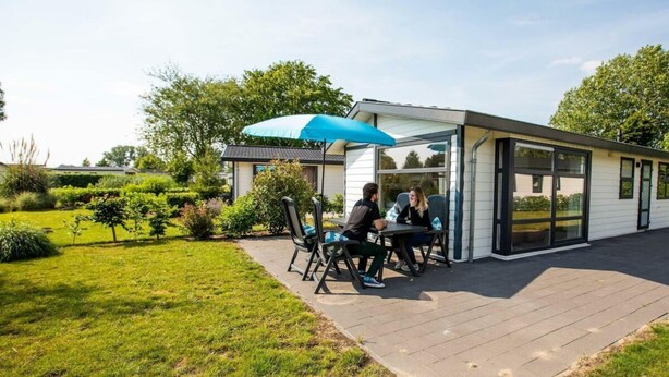 Un área de estar moderna al aire libre con una mesa y sillas bajo un paraguas azul, contigua a un edificio con amplias ventanas.