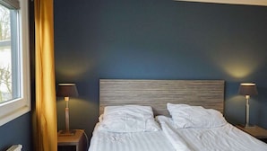 Cama doble con sábanas blancas, dos mesitas de noche con lámparas, una ventana con cortinas y una pared azul oscuro.