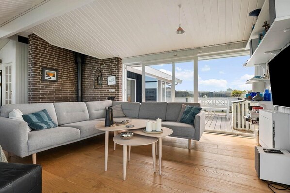 A modern living room with a grey sofa, a small round table, and a kitchen area with a white refrigerator. - LN2066 Hjørring - Slettestrandvej 3 (Lønstrup)