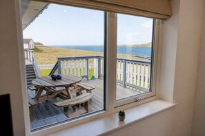 Et værelse med et stort vindue der kigger ud på en terrasse med et træbord og bænke, en lille potteplante og udsigt til havet og landskabet i det fjerne.