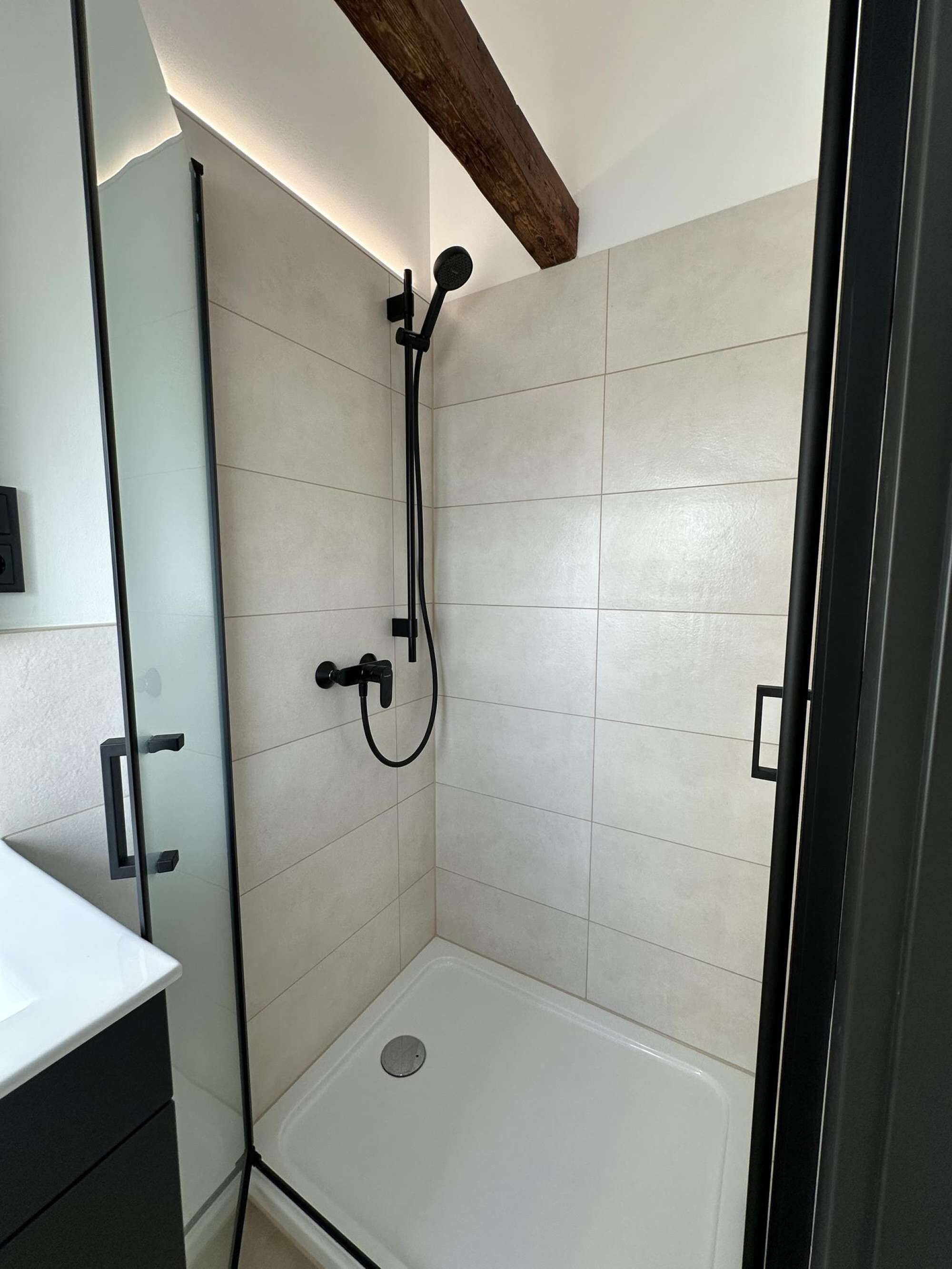 Une douche avec une porte en verre, des murs recouverts de carreaux blancs et un plafond avec une poutre en bois.