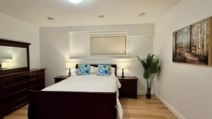 A bedroom with a bed, two bedside tables with lamps, a large mirror, a window with blinds, a potted plant, and a painting on the wall.