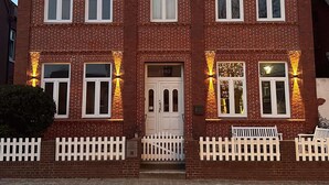 A two-story brick building with multiple windows, a white door, and a small porch with benches. - Stylish OSTEREMS Apartment in STRAND HAUS 10 (Borkum)