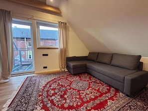 A room with a grey sectional sofa, a red patterned rug, a large window with curtains, and a view of a brick building outside. - Stylish OSTEREMS Apartment in STRAND HAUS 10 (Borkum)