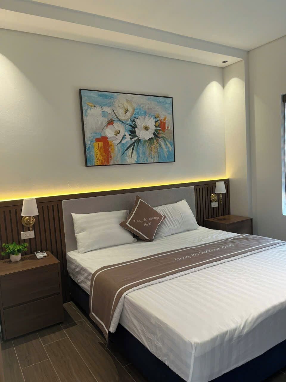 Une chambre d’hôtel avec un grand lit, une table de chevet avec une lampe, un tableau accroché au mur et une petite table de chevet ornée d’une plante.