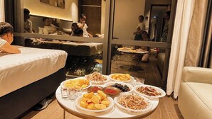 Una habitación de hotel con una cama, una mesa de comedor y diversos alimentos, y un espejo que refleja la habitación.