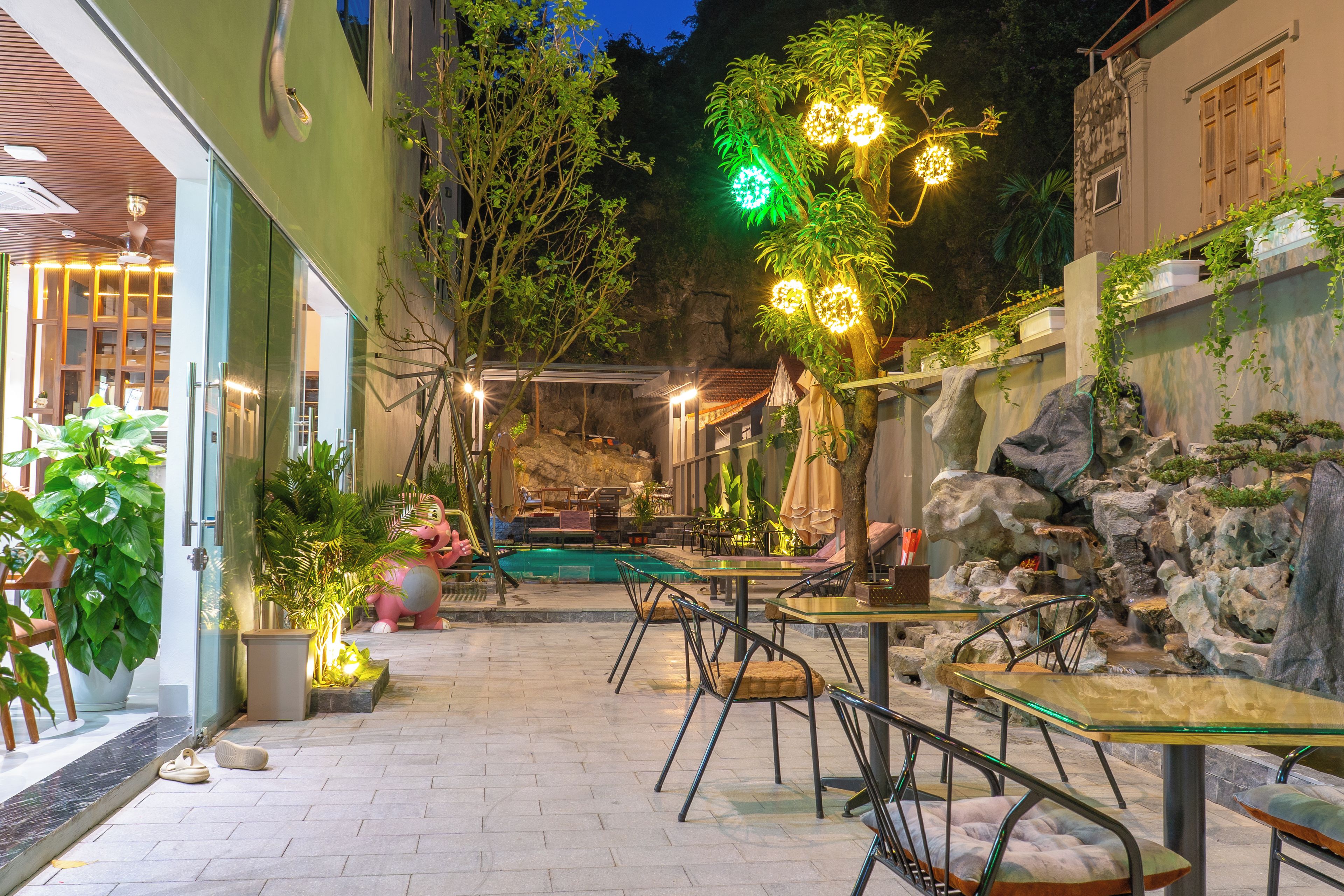 An outdoor patio area with tables and chairs, surrounded by greenery and a small pool.