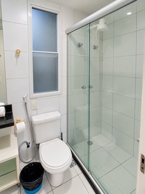 A bathroom with a glass-enclosed shower, a toilet, a window, and a trash bin. - Beach Village sea View 2 Bedrooms (Fortaleza)