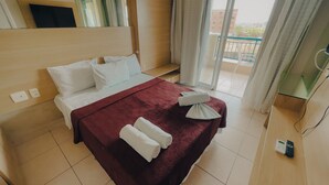 A hotel room with a bed, a red bedspread, white pillows, a white towel, a bedside table, a mirror, and a view of the outside. - Beach Village sea View 2 Bedrooms (Fortaleza)