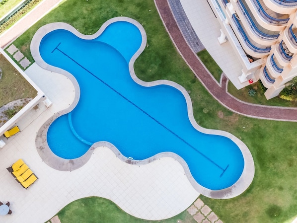 An aerial view of a swimming pool with a unique wavy design, surrounded by a paved area with yellow chairs and a white building with blue balconies. - Beach Village sea View 2 Bedrooms (Fortaleza)