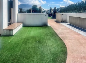 Eine Dachterrasse mit künstlichem Rasen, Betonpflanzgefäßen und Blick auf Berg und Stadt.