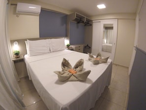 Ein ordentlich bezogenes Bett mit Handtüchern, die zu einem Blumenmuster arrangiert sind, ein Nachttisch mit einer Lampe, ein Kleiderständer und ein Spiegel an der Wand.