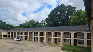 A multi-story motel with a parking lot, a white car parked, and a dog near the entrance. - Cajun Inn  (Shreveport)