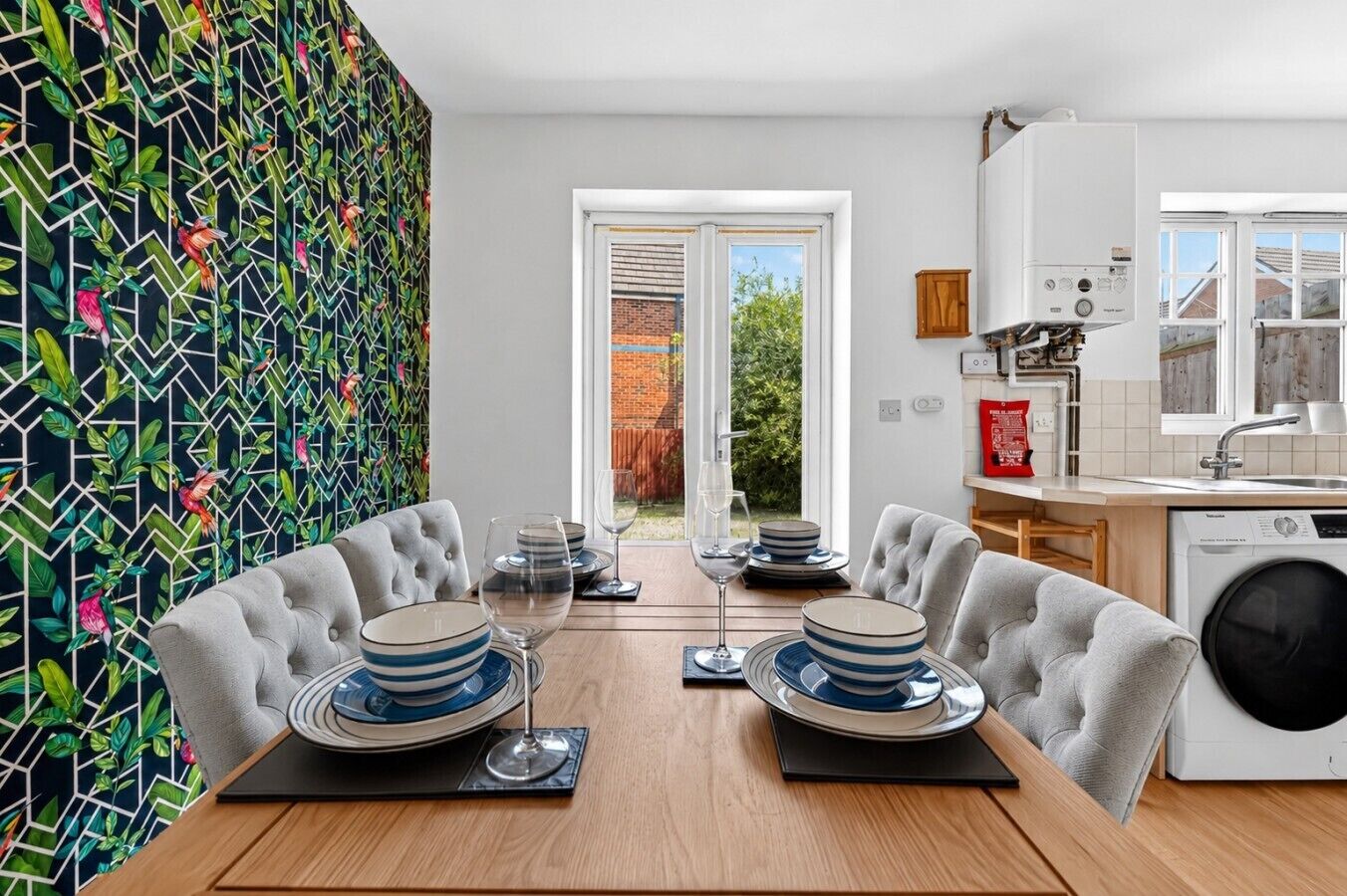 Un comedor con una mesa dispuesta para dos, una cocina con lavadora y vistas al exterior a través de un ventanal.