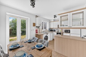 Cozinha moderna com armários brancos, área de jantar com mesa de madeira posta para quatro pessoas e vista para o exterior por uma porta deslizante.