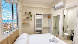 A hotel room with a bed, a wardrobe, a shelf with decorative items, and a view of the sea. - Apartamento Novo 2 Quartos e Vista mar 1001b (Fortaleza)