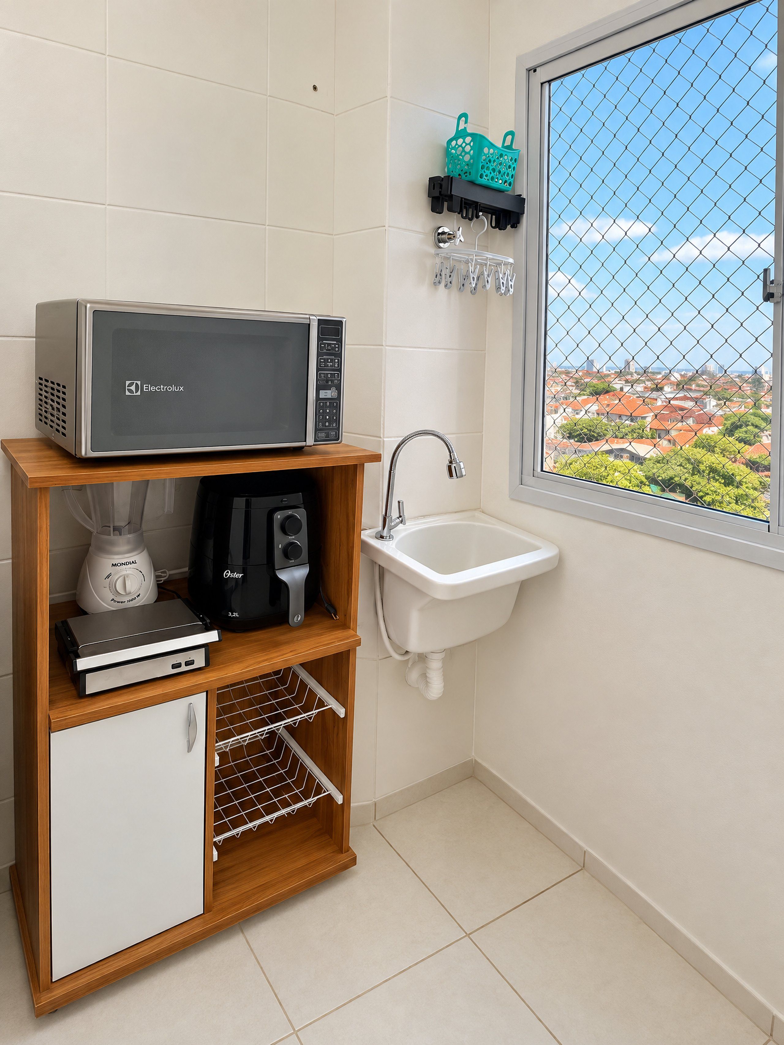 Una cocina con microondas, licuadora y cafetera sobre una repisa de madera, un lavamanos blanco y una ventana con vista a edificios.