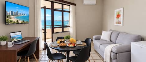 A modern living room with a dining area, a laptop on a desk, and a view of the sea through large windows.