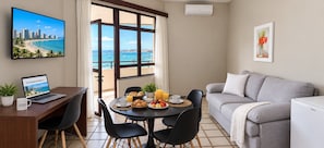 A modern living room with a dining area, a laptop on a desk, and a view of the sea through large windows.